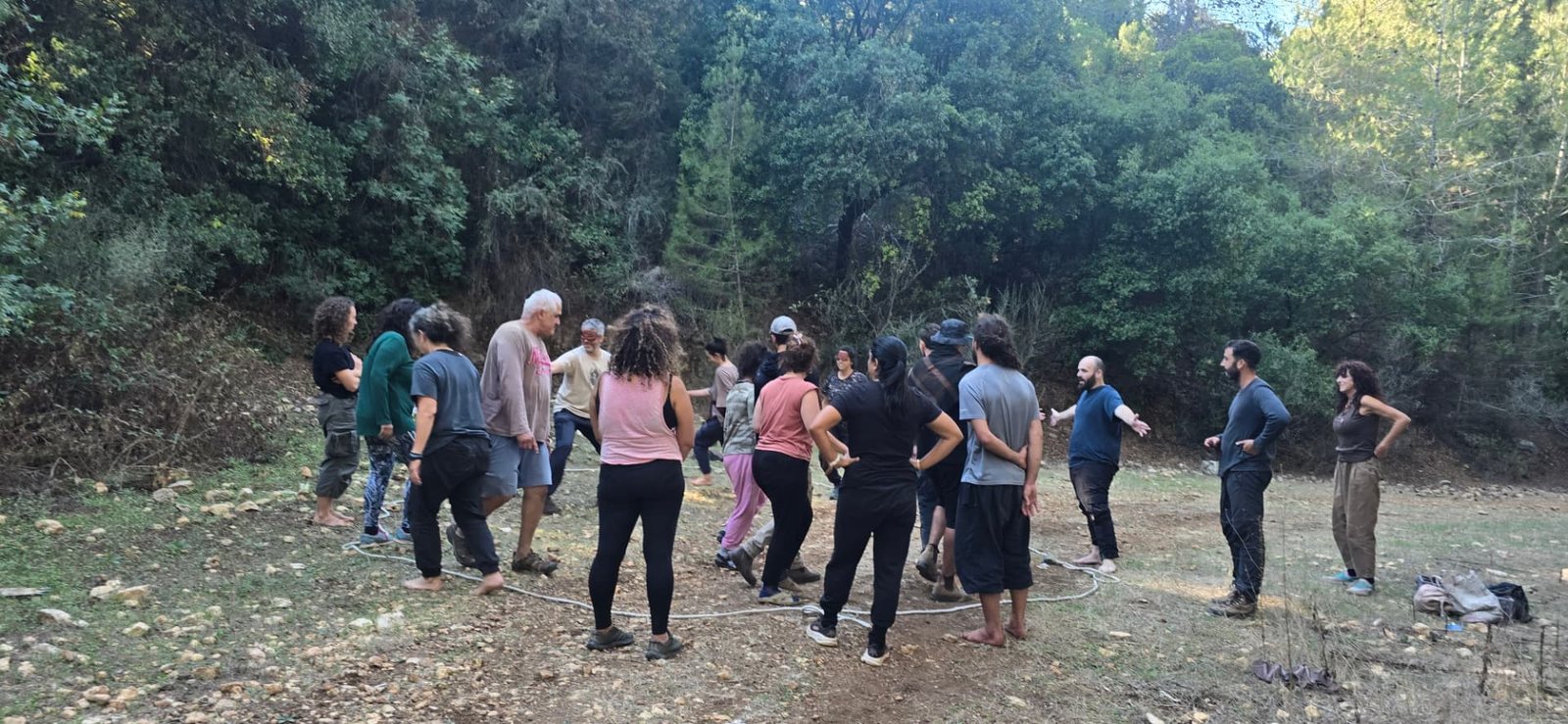 קבוצת משתתפים עומדת במעגל בקרחת יער, בין עצים ואור אחר צהריים